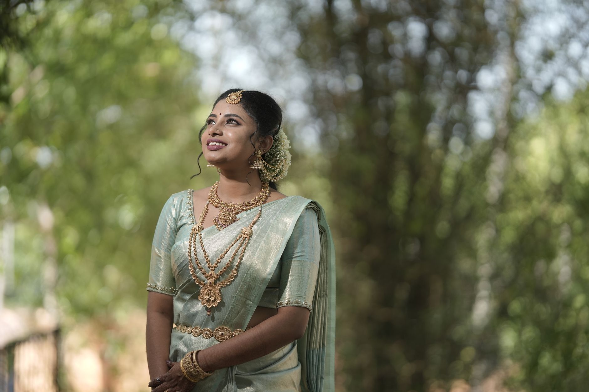 A woman in traditional Indian attire with jewelry, posing outdoors with lush green background.